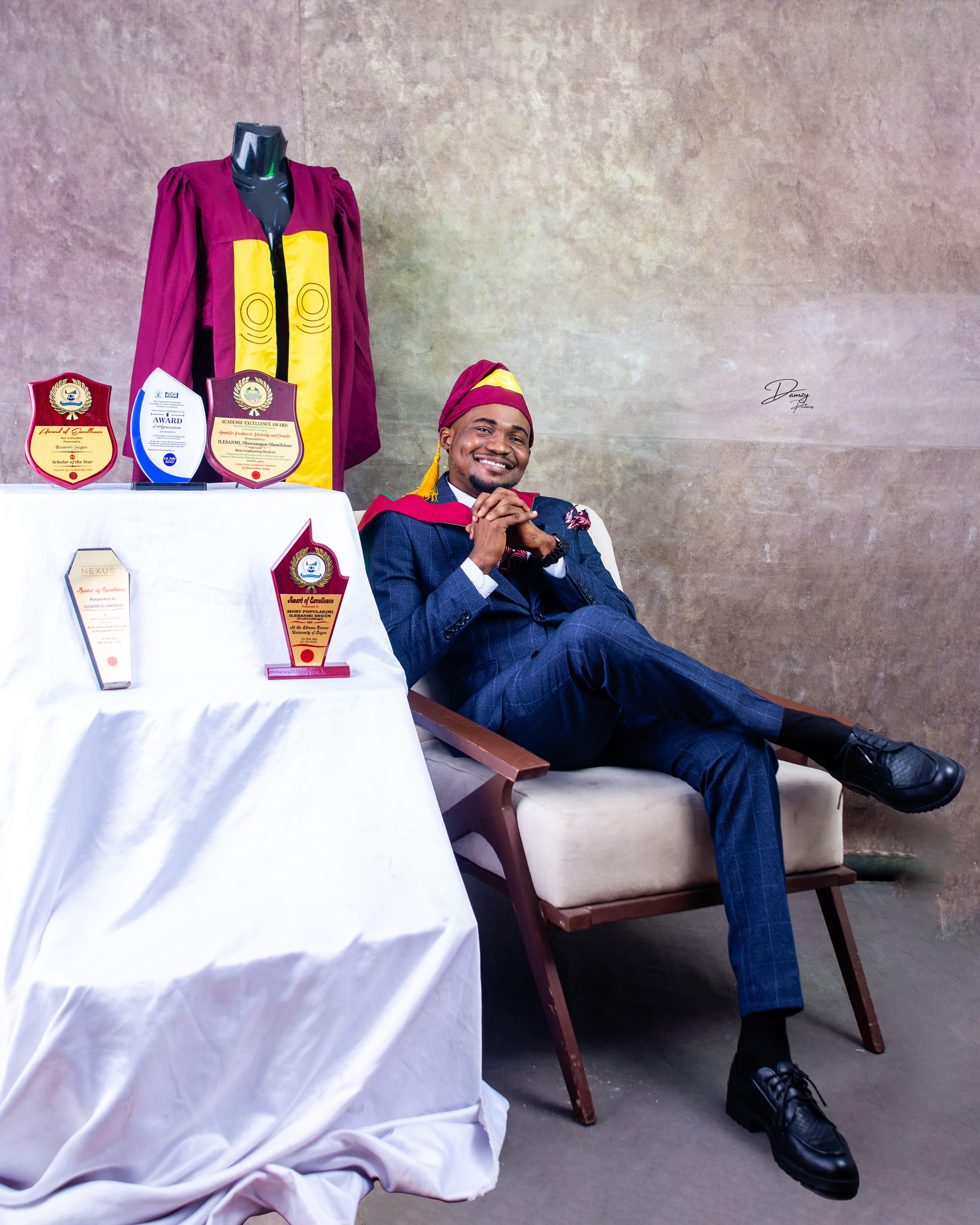 Graduate in a navy suit seated beside a convocation gown and award plaques