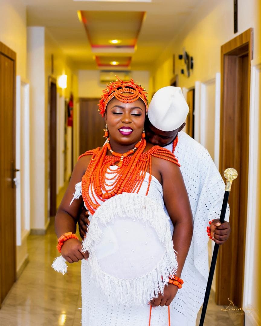Bride in coral beads and white lace beside her groom holding a cane in a hotel corridor