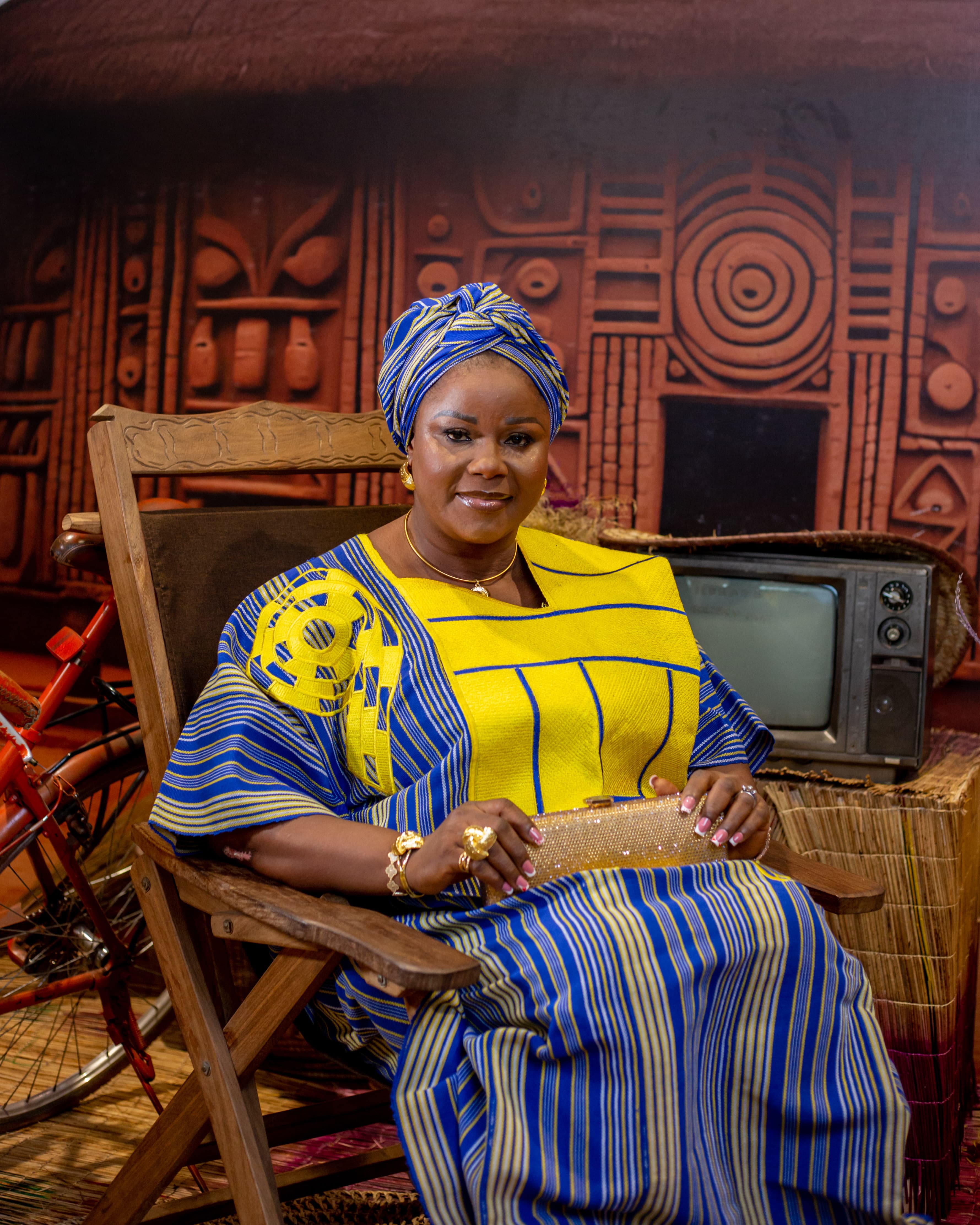 Studio portrait of a woman in yellow and blue traditional attire seated on a carved chair